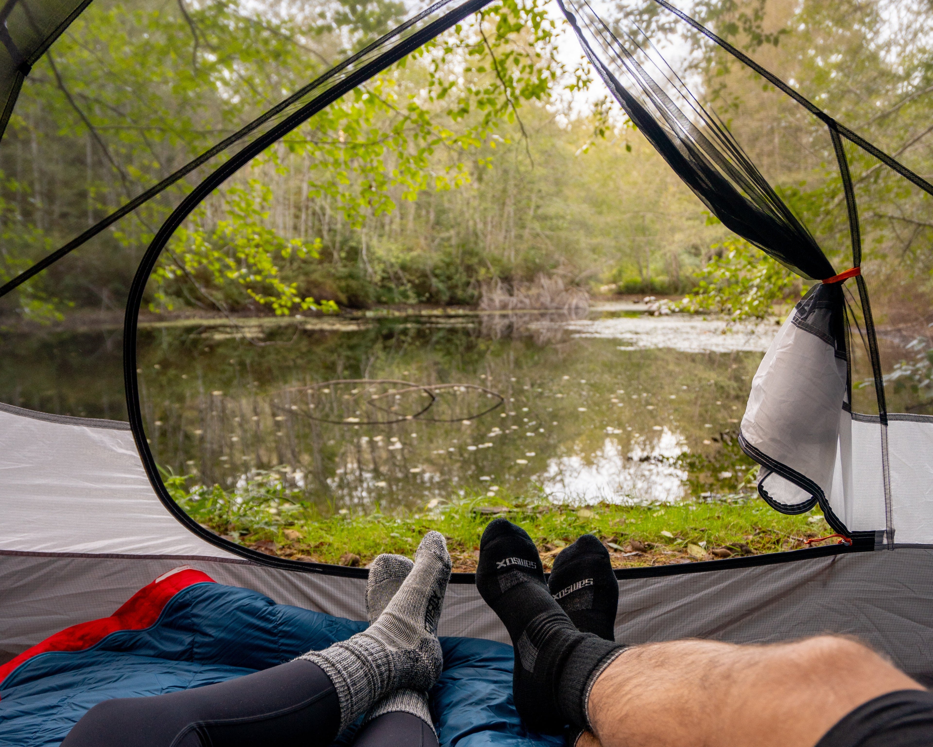 Two people lying inside a tent overlooking a serene lake surrounded by trees.